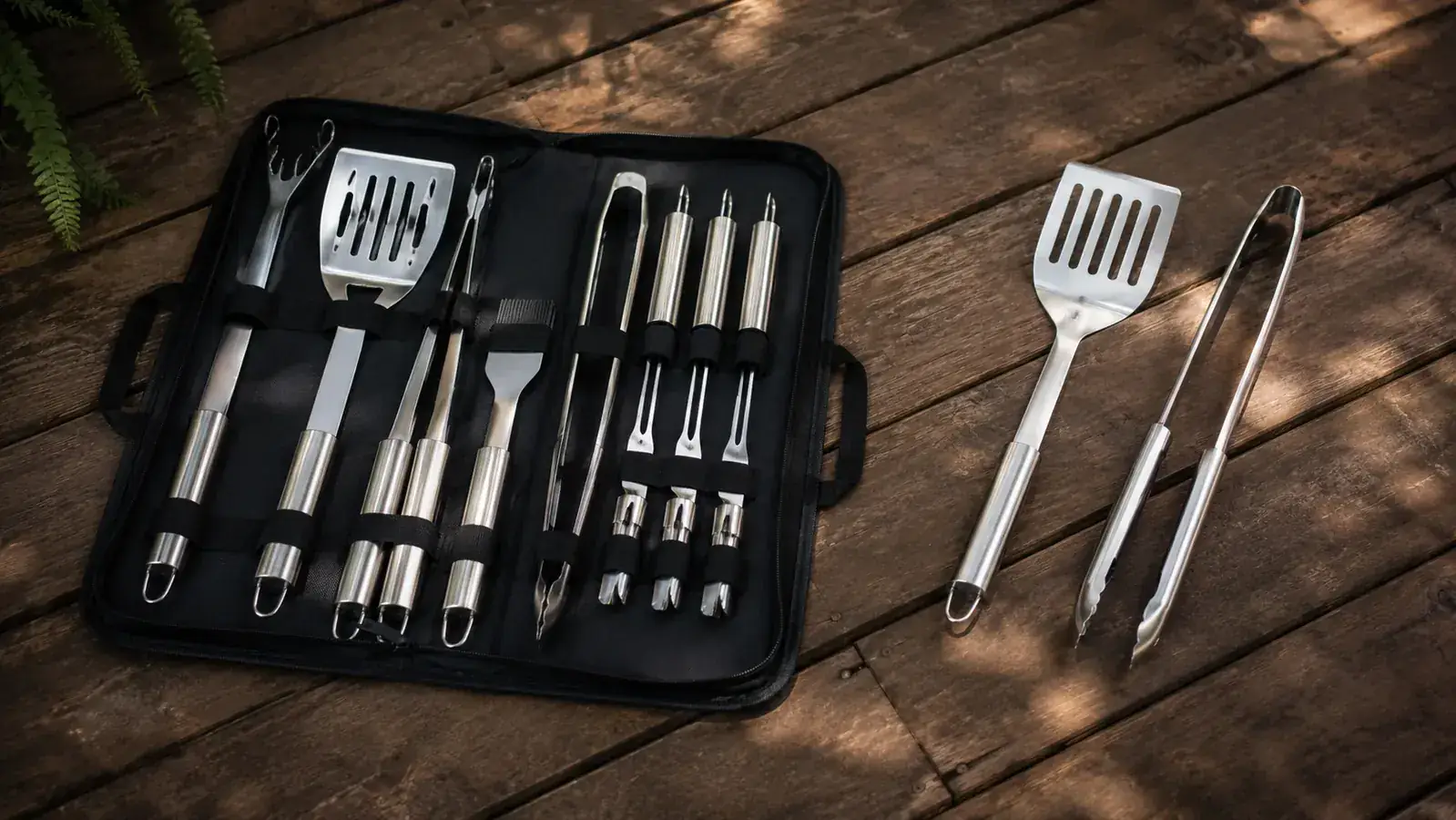 ROMANTICIST 23-piece BBQ tool set laid out on a weathered wood picnic table next to a charcoal grill, tools fanned out of the carry case