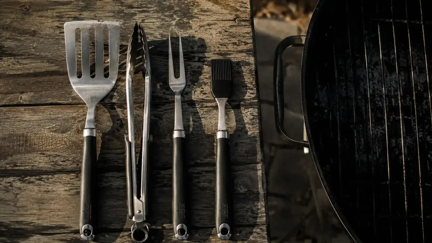 ROMANTICIST 23-piece BBQ grill tool set laid out on a weathered wood picnic table beside a charcoal kettle grill