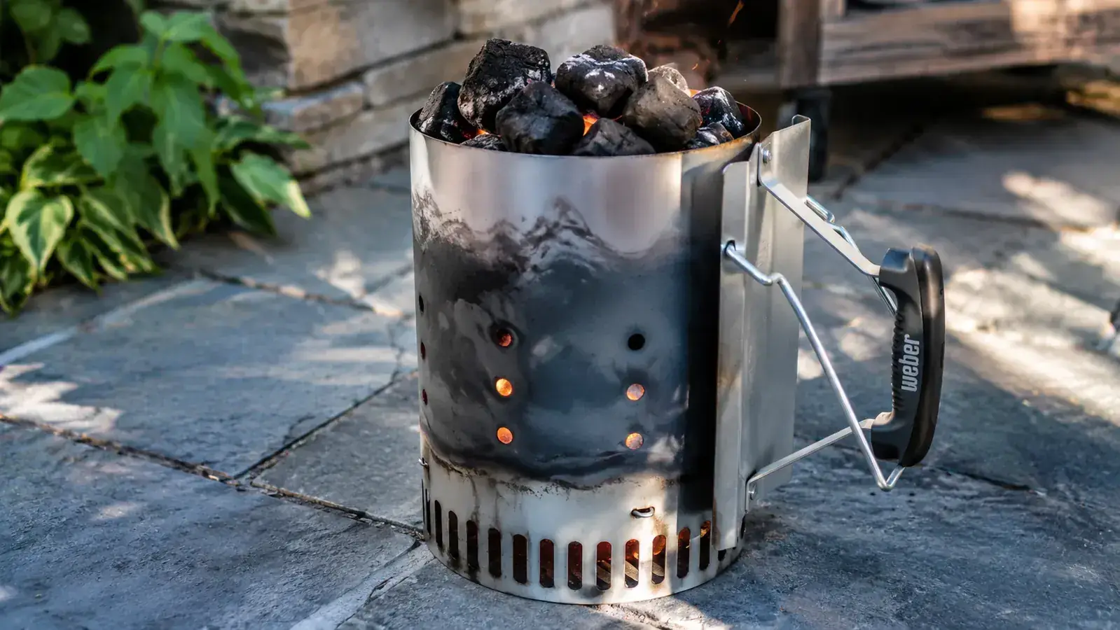 Weber Rapidfire Chimney Starter sitting on a concrete patio with lit coals glowing orange at the top, smoke rising against a late-afternoon sky