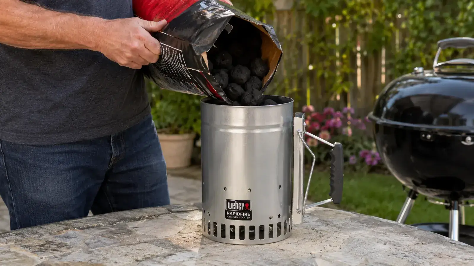 Close-up of a Weber chimney starter being held over a charcoal grill, ready to pour lit coals
